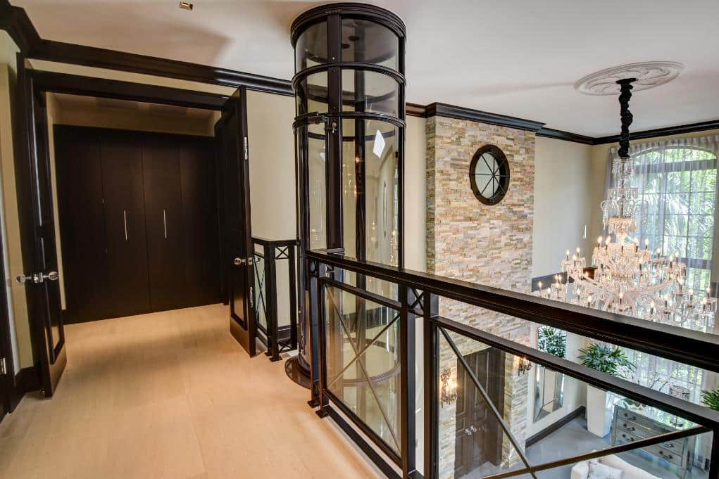 A stylish hallway with a glass railing, leading to a room with a chandelier and stone wall. Black doors and elegant decor are visible.