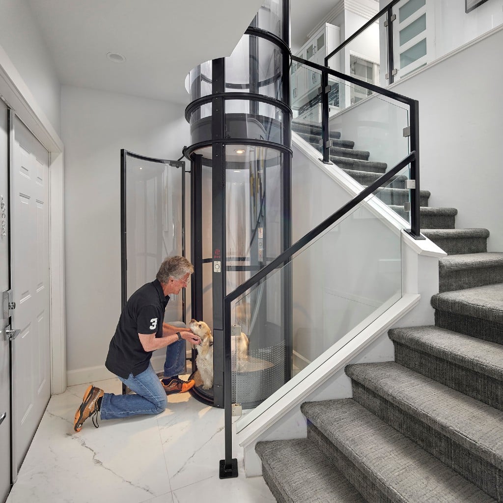 A man kneels beside a dog at a glass elevator in a modern home, with stairs visible in the background.